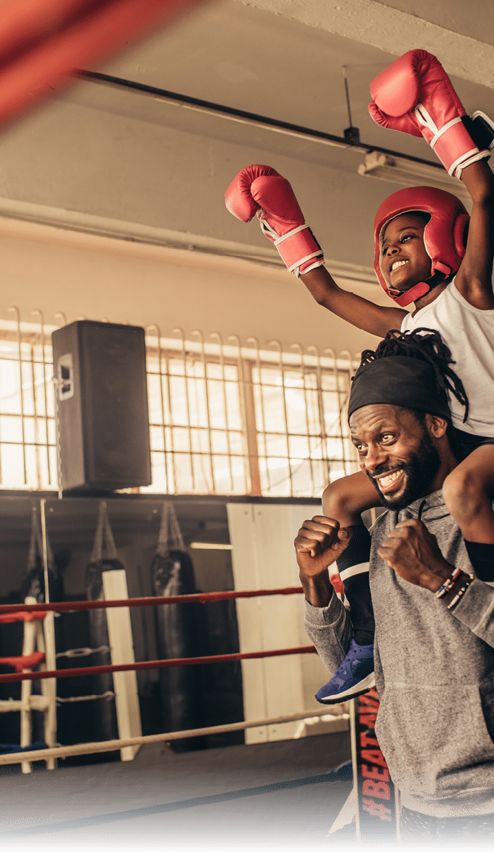 Trainer carrying a boxer kid on shoulders celebrating a win  Kid in boxing gear sitting on shoulder of coach with raised hands 